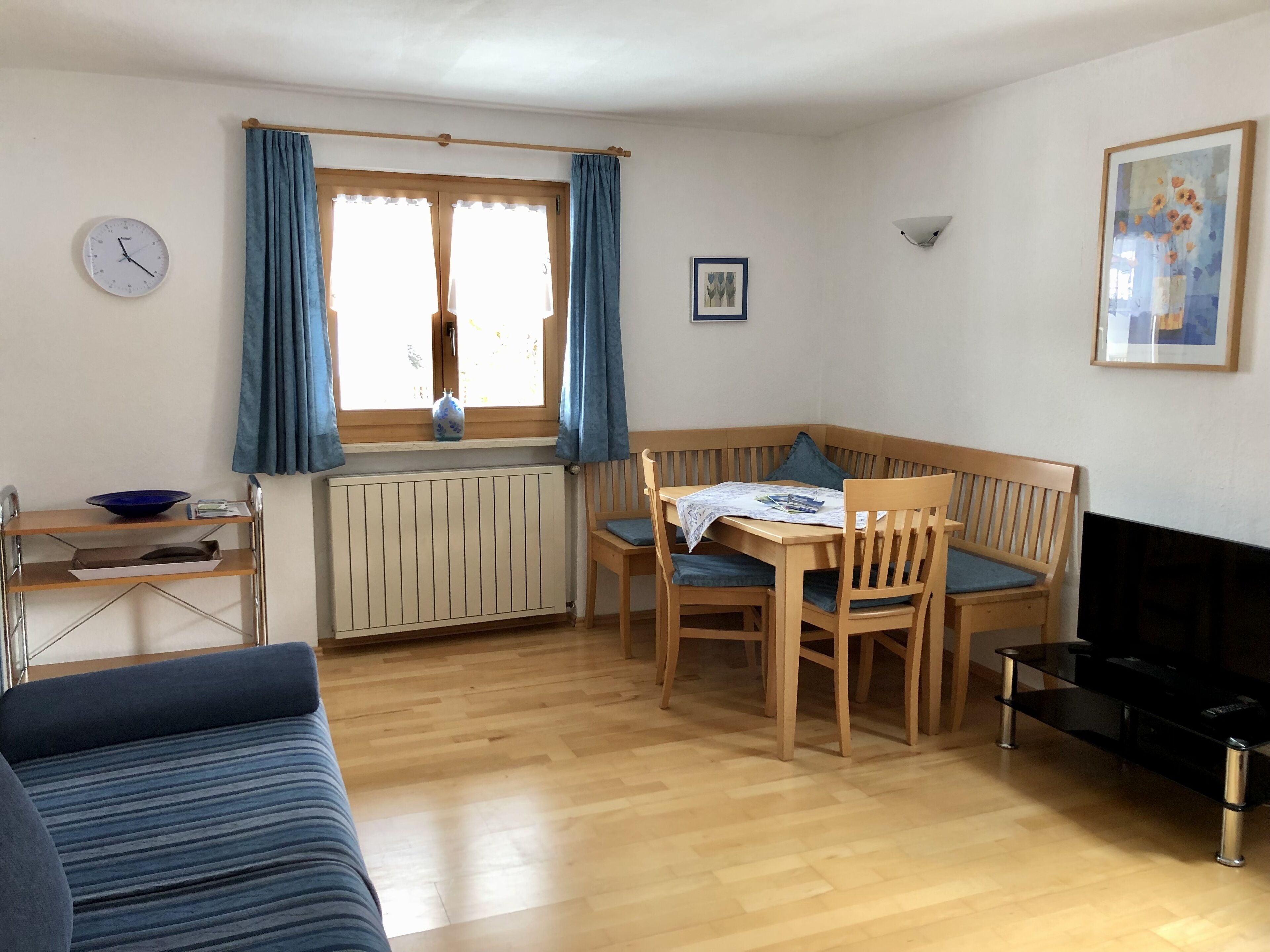 A cozy room with a wooden dining table, two chairs, a blue sofa, a small TV, and a window with curtains.