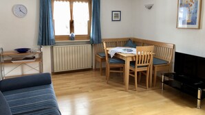 A cozy room with a wooden dining table, two chairs, a blue sofa, a small TV, and a window with curtains. - (2) Zwei-Raum-Ferienwohnung 40qm, 1-4 Personen, Extra-Schalfzimmer, Wohnküche, große Balkonterrasse (Reit im Winkl)
