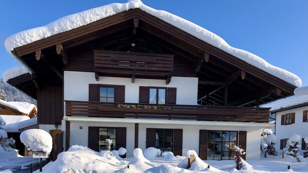 A two-story chalet with a sloped roof covered in snow, wooden exterior, and large windows. - (2) Zwei-Raum-Ferienwohnung 40qm, 1-4 Personen, Extra-Schalfzimmer, Wohnküche, große Balkonterrasse (Reit im Winkl)