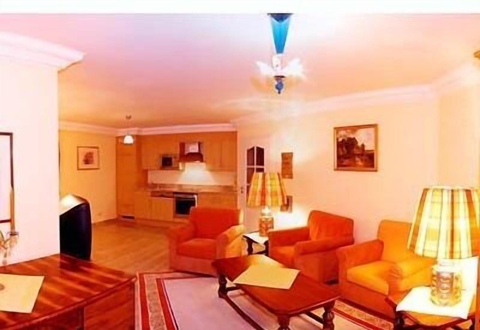 Ein Wohnzimmer mit orangefarbenen Sofas, einem Couchtisch und einem Fernseher. Im Hintergrund ist eine Küche mit einem Herd und Schränken zu sehen.