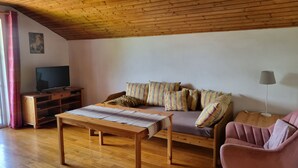 Una sala de estar acogedora con techo de madera, un sofá con cojines a rayas, un televisor sobre un mueble de madera, una mesita y una silla rosa.