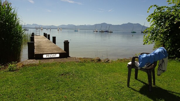 Un muelle privado que da a aguas tranquilas con veleros, montañas al fondo y una silla con una chaqueta.