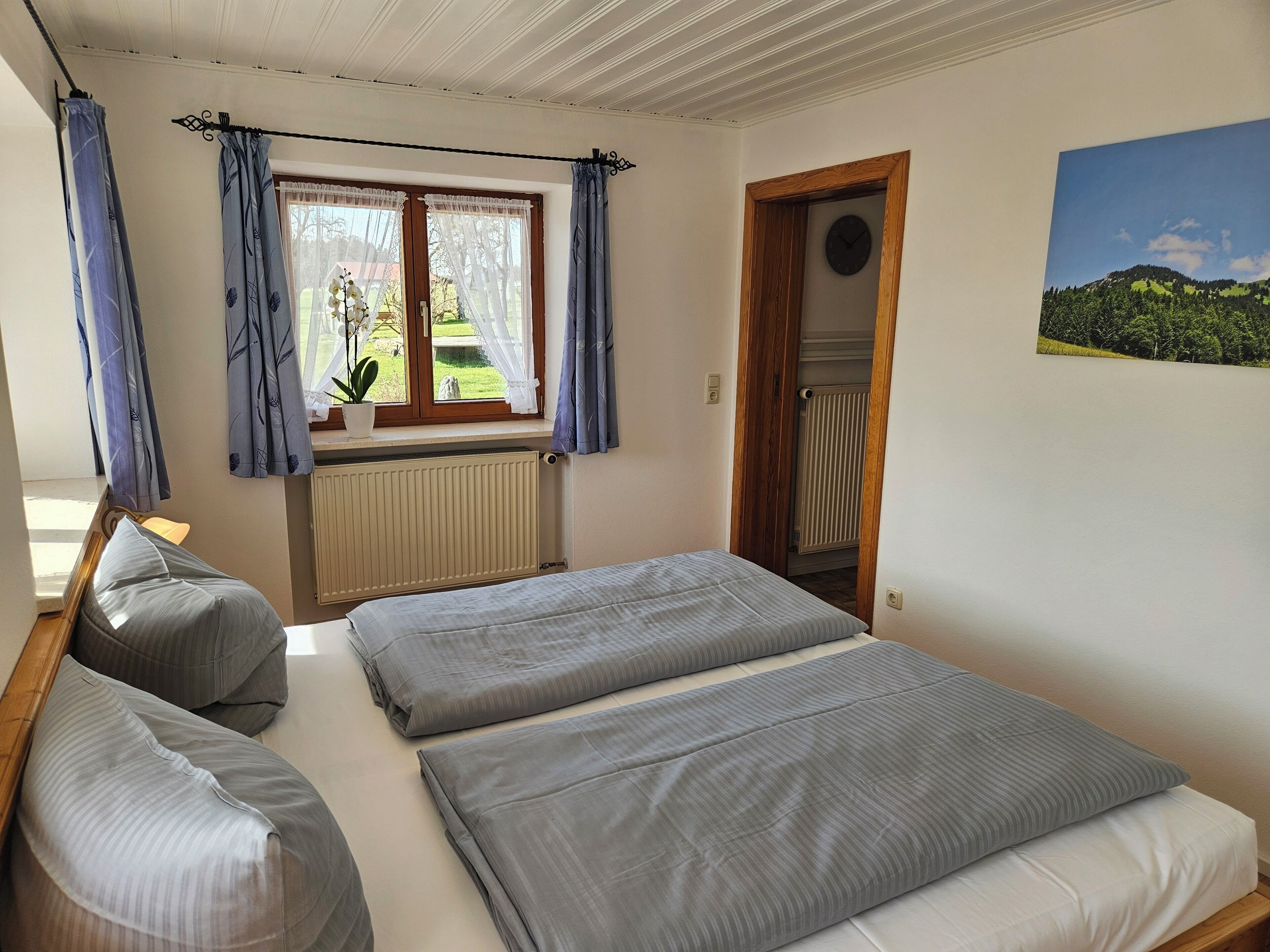 A bedroom with a bed, a window with curtains, a radiator, and a door leading to another room.