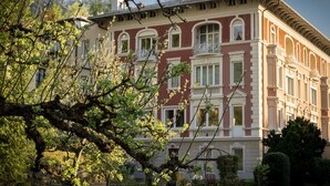 Ein großes, historisches Gebäude mit zahlreichen Fenstern und Balkonen, umgeben von üppiger Bepflanzung und einem gepflegten Rasen.