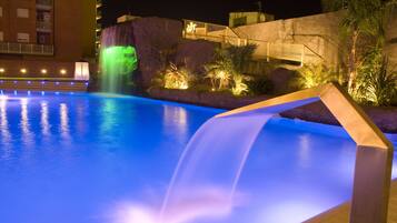 Un área de piscina nocturna con una cascada, un muro de piedra y una escalera que conduce a un nivel superior.