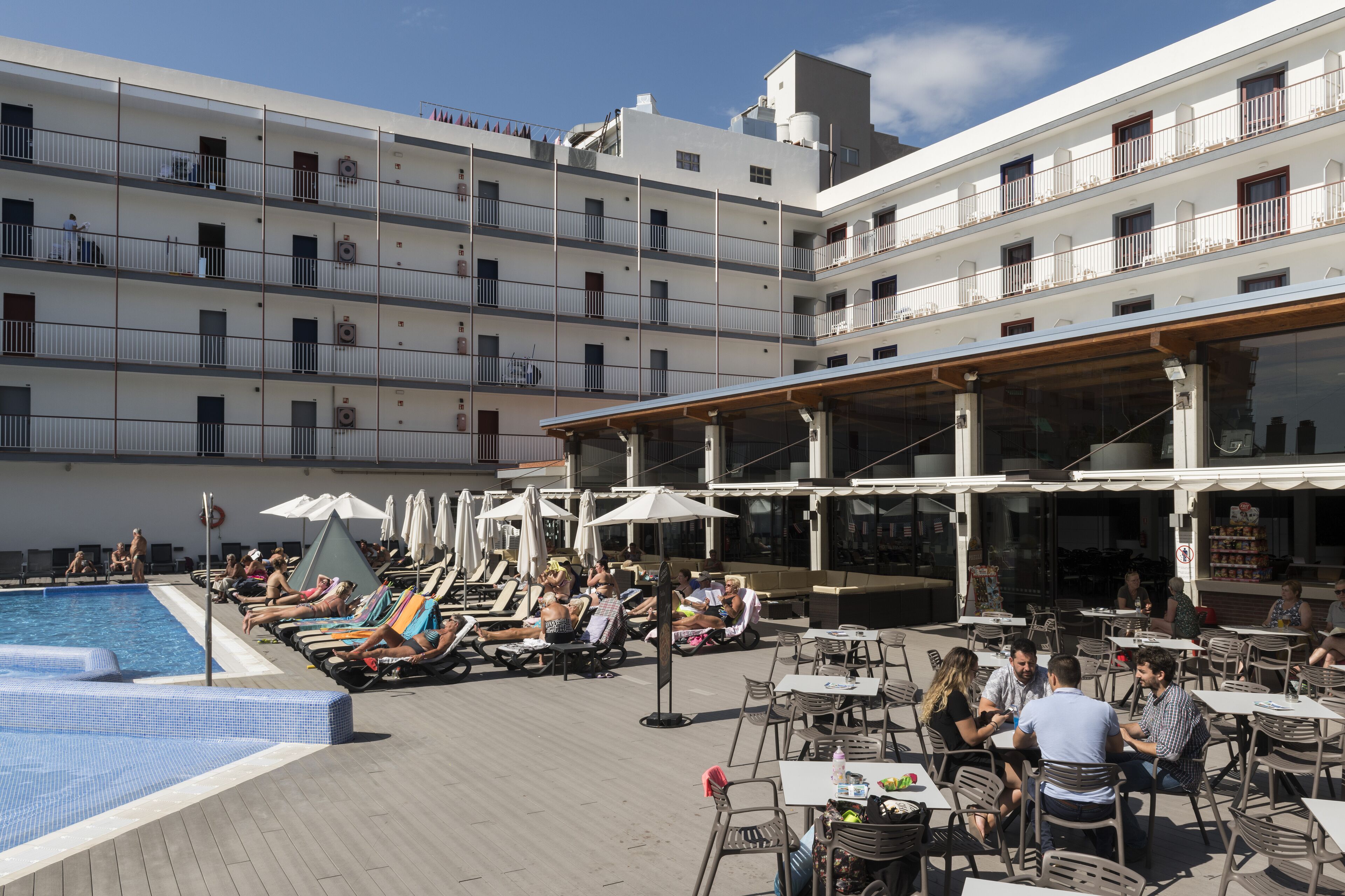 A rooftop pool area with lounge chairs and umbrellas, surrounded by outdoor seating and a building with multiple balconies.