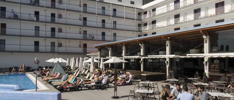 A rooftop pool area with lounge chairs and umbrellas, surrounded by outdoor seating and a building with multiple balconies.