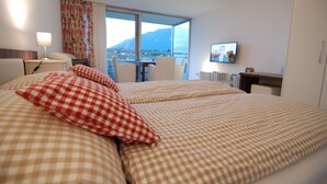 Hotellihuone, jossa on suuri sänky, työpöytä tuolilla, televisio ja ikkunasta avautuva näkymä vuorille.