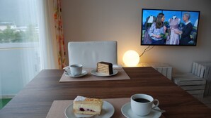 Hotellihuoneessa on puinen pöytä, jonka päällä on viipale kakkua, kuppi kahvia ja televisio, josta näkyy jokin ohjelma.