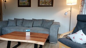Een moderne woonkamer met een grijze bank, een houten salontafel, een blauwe fauteuil, en twee ingelijste schilderijen aan de muur.