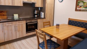 Een compacte keuken met houten kasten, een inbouwoven en een koffiezetapparaat. Er is een eettafel met stoelen en een klok aan de muur.