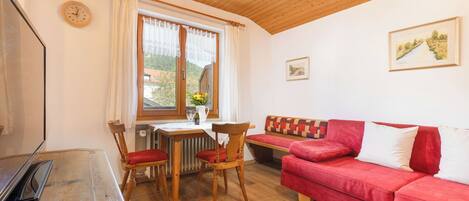 A cozy room with a wooden ceiling, a red sofa, a wooden table with chairs, and a window with curtains.