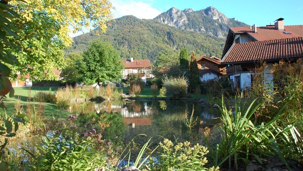 A serene lakeside scene with traditional buildings, a mountain backdrop, and lush greenery.