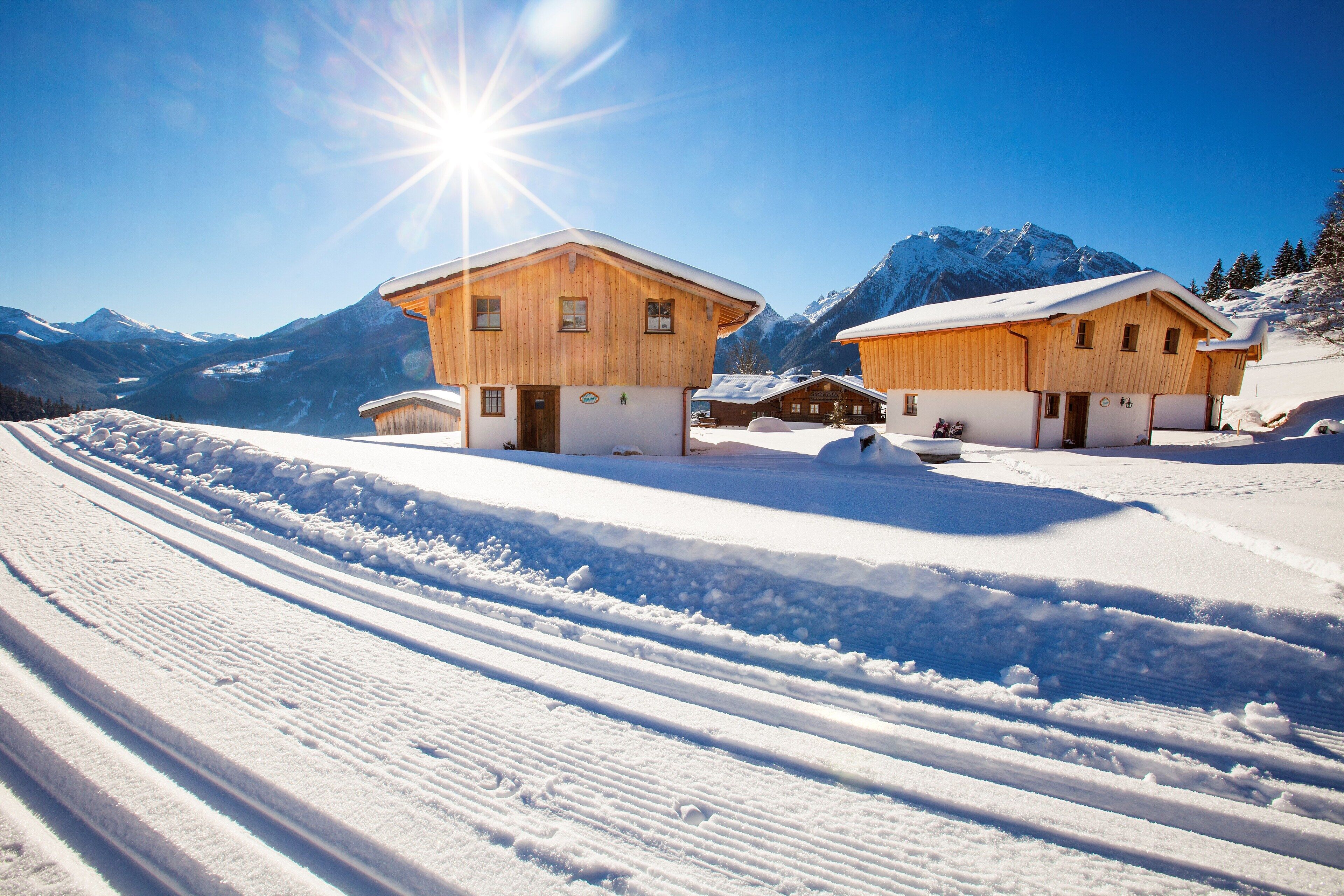 A wooden chalet with a snow-covered roof, set against a backdrop of mountains and a clear sky.