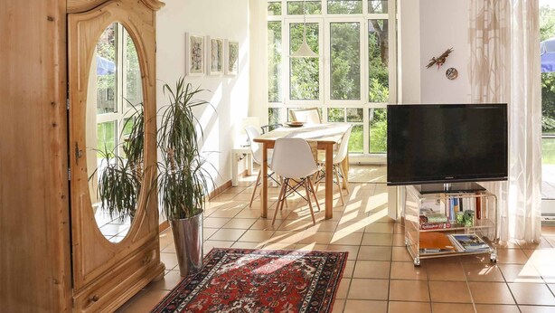 Un salon avec un meuble en bois, un téléviseur posé sur un meuble, une table à manger avec des chaises et une grande fenêtre avec des rideaux.