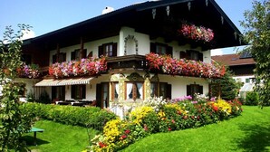 Un bâtiment de style alpin traditionnel, doté d’un balcon orné de fleurs aux couleurs vives, d’un jardin bien entretenu et surplombant un ciel d’un bleu éclatant.