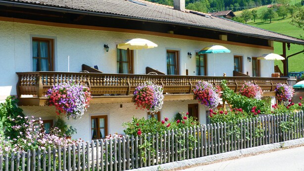 A two-story building with a balcony, a wooden fence, and hanging flower baskets. - Ferienwohnung I 45 qm°, zwei Schlafzimmer, Südbalkon und ebenerdigem Eingang° (Aschau im Chiemgau)
