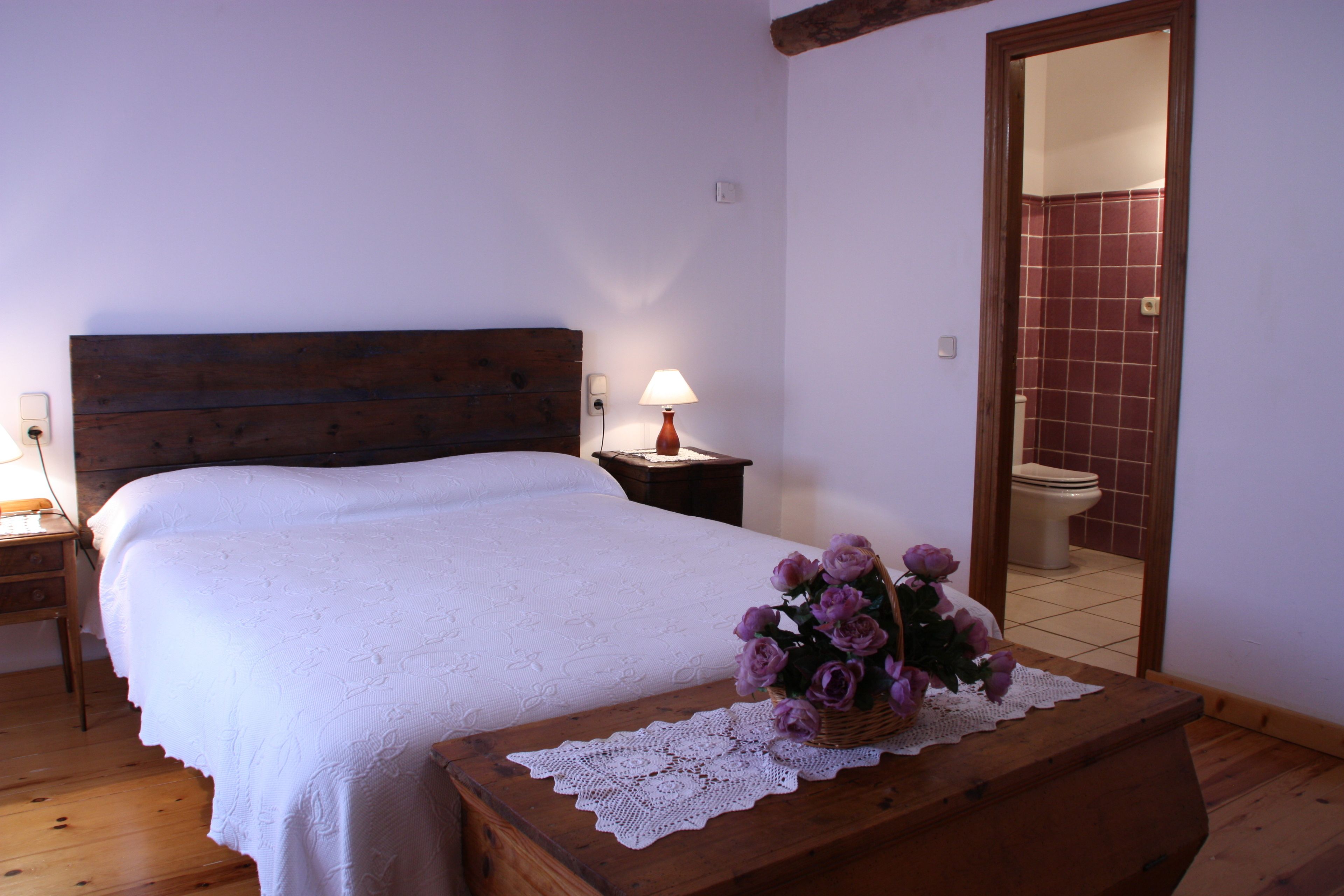 Un dormitorio con cabecera de madera, cubrecama blanco, mesita de noche con lámpara y mesita auxiliar de madera.