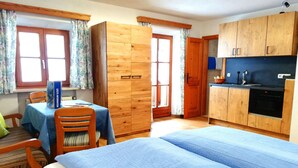 Une pièce comprenant un lit, une table avec des chaises, une armoire en bois, un coin cuisine avec un évier et une cuisinière, et une fenêtre avec des rideaux.
