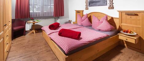 Een houten bed met rood-witte geruit beddengoed, een houten nachtkastje met een schaal fruit, een houten kledingkast, een rode stoel en een raam met rode gordijnen.