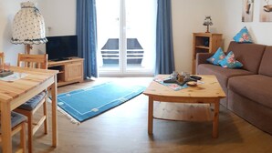 Un salon avec une table en bois, un canapé et un téléviseur. Il y a un balcon avec des rideaux bleus.