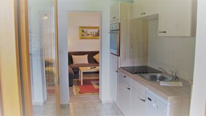 A compact kitchen with a built-in oven, a sink, and a dishwasher. There is a small seating area with a sofa and a painting on the wall. - Ferienwohnung Grünstein, 1 bis 2 Personen, 54 qm, 1 Schlafzimmer, extra Küche, Balkon (Schönau am Königssee)