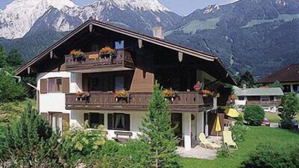 Un chalet doté d’un balcon, entouré d’une végétation luxuriante et avec des montagnes en toile de fond.