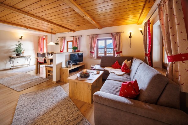 A cozy living room with a wooden ceiling, a sofa with red cushions, a television, and a small table with a decorative bowl. - Apartment Alm Stüberl, 65 square meters, balcony, 2 sep. Bedroom, max. 4 pers. (Ruhpolding)