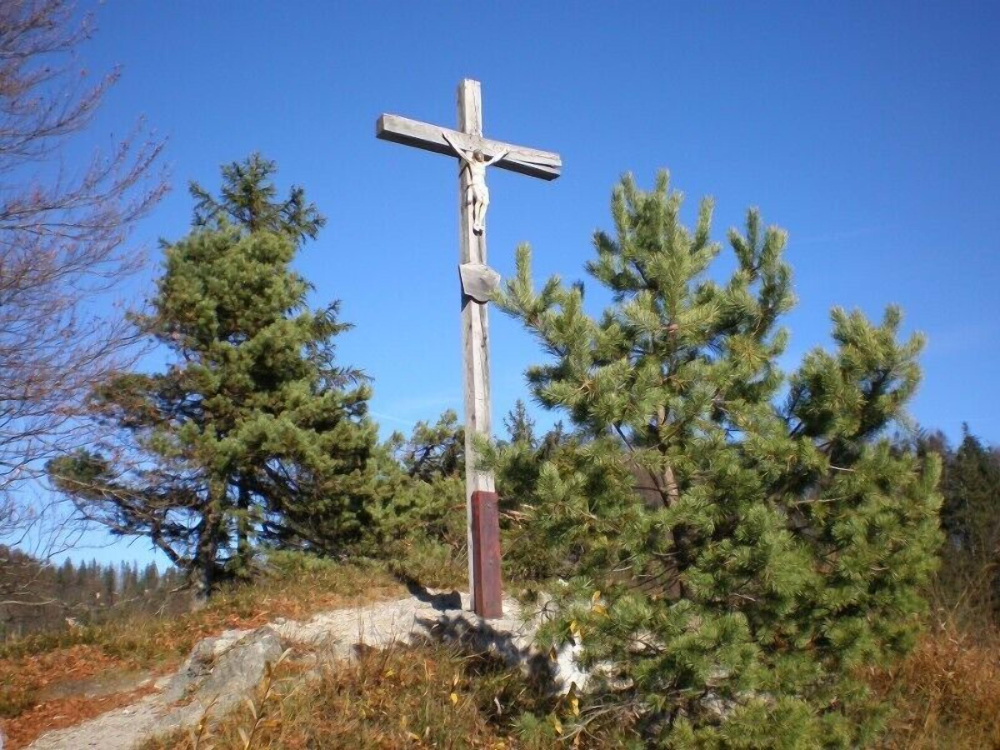 Una cruz de madera con la figura de la crucifixión, rodeada de pinos y un cielo despejado.