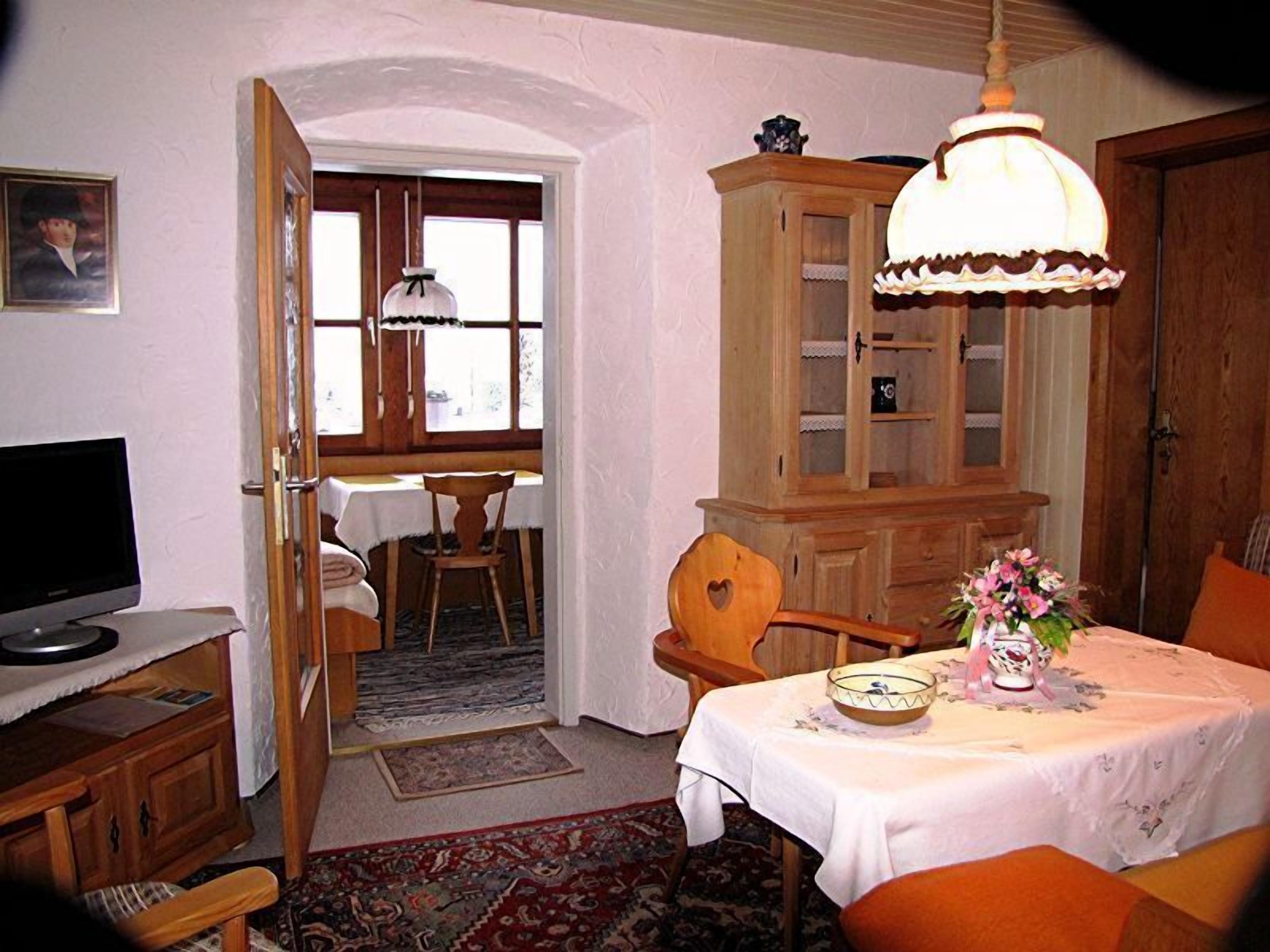 Um quarto com uma mesa de jantar posta para dois, uma televisão em um suporte, um armário com portas de vidro e uma janela grande com cortinas.