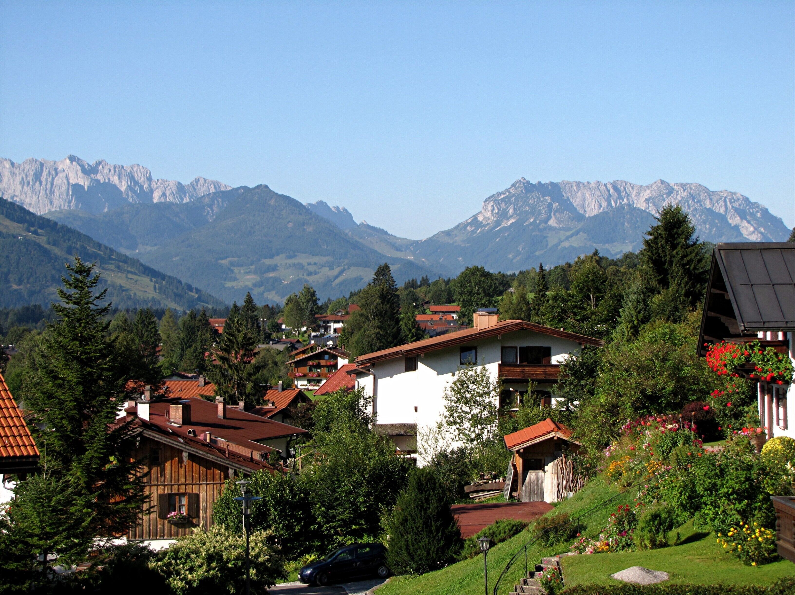 Un village de montagne avec des maisons traditionnelles en bois, une voiture garée devant, et une vue sur les montagnes en arrière-plan.