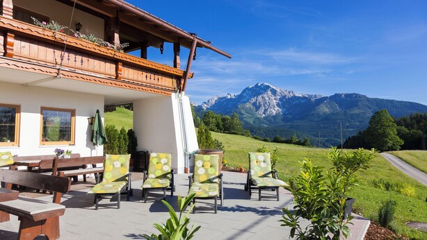 An outdoor seating area with wooden benches and chairs, a balcony with flowers, and a mountain view.