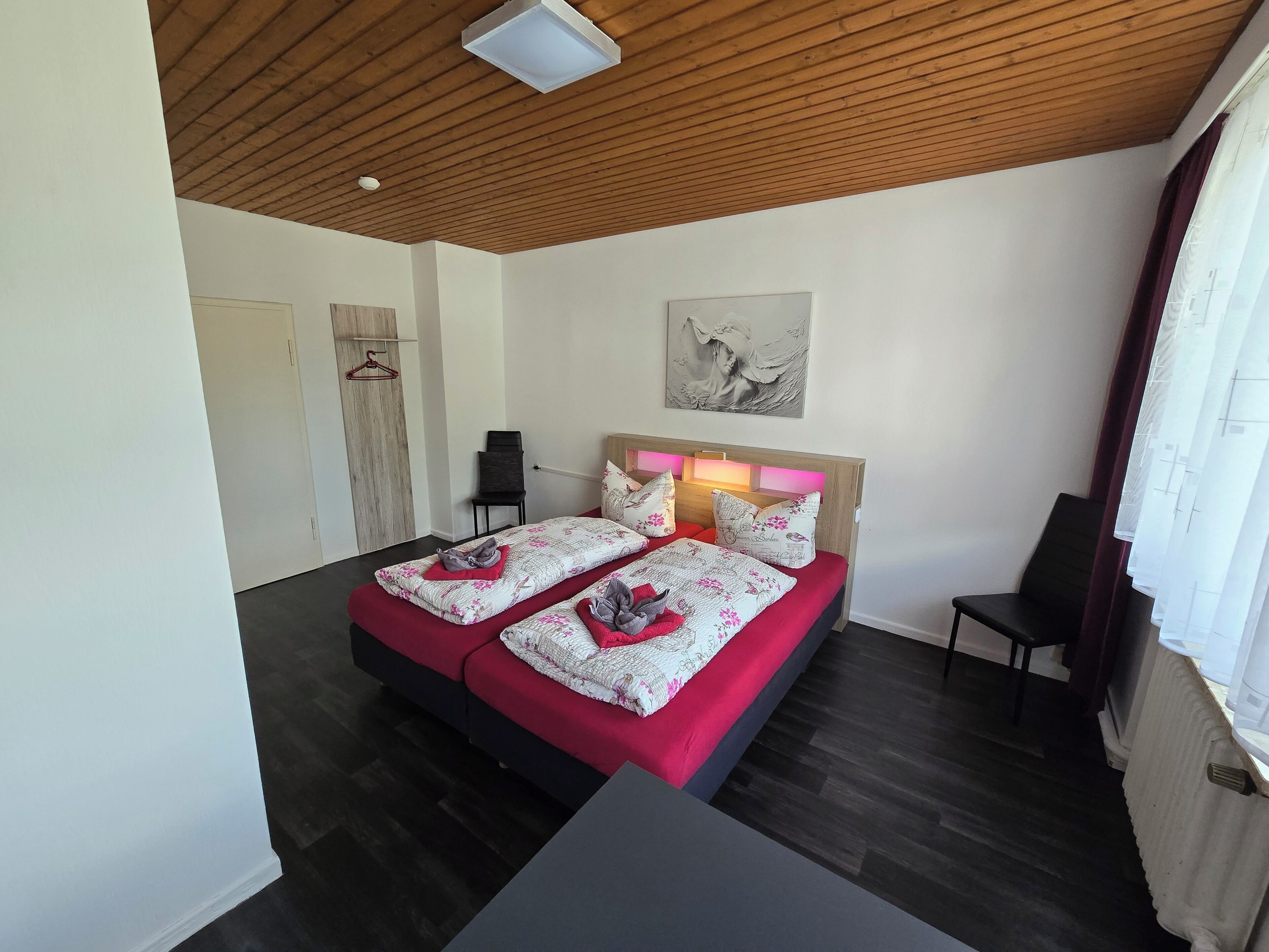Une chambre avec un plafond en bois, un lit avec une literie rouge et blanche, une chaise noire et un tableau accroché au mur.