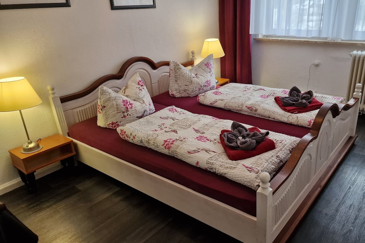 A double bed with floral bedding and towels, a bedside table with a lamp, a radiator, and a window with curtains.