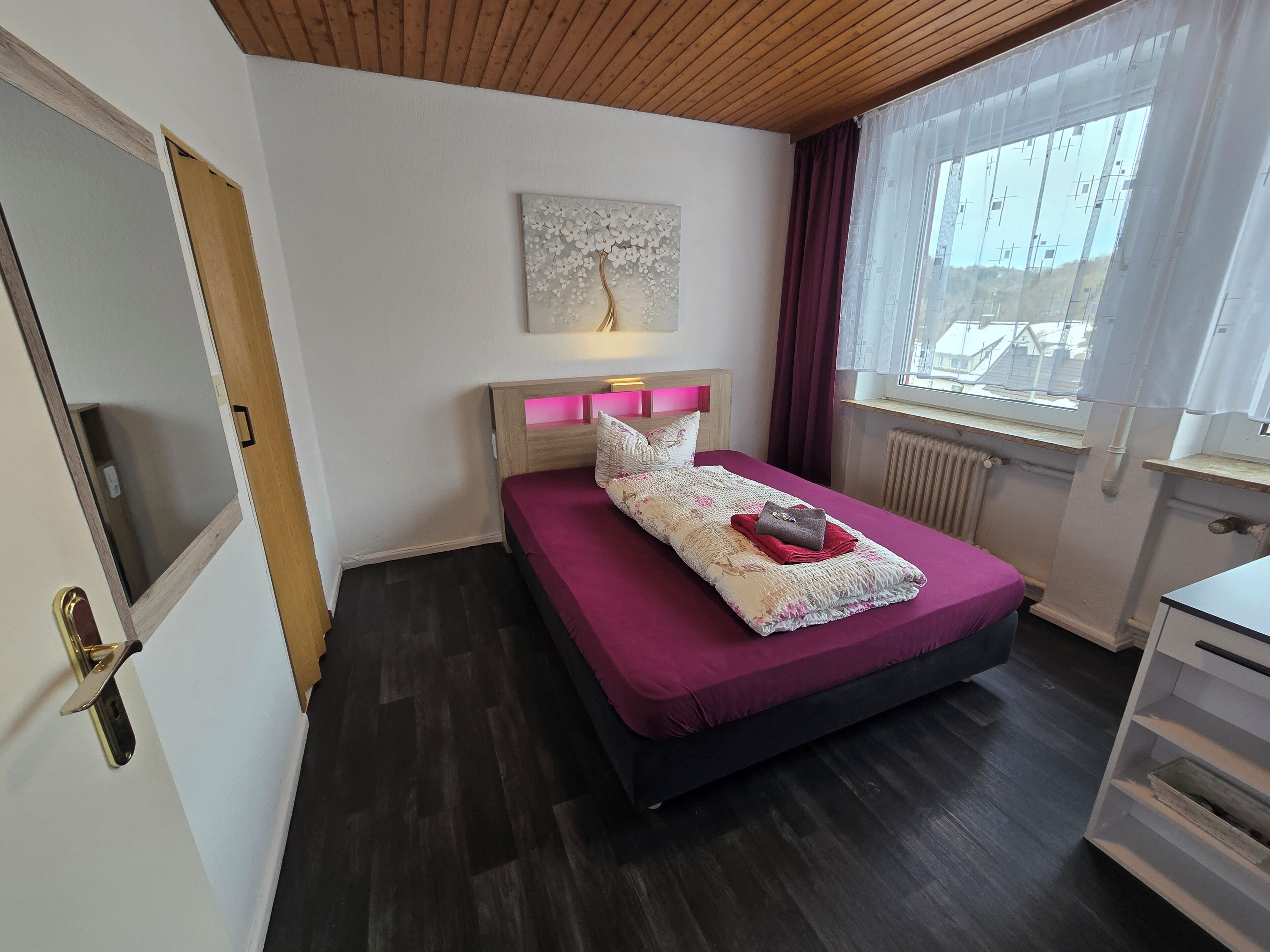 Een slaapkamer met een bed, een raam met gordijnen, een radiator en een houten plafond.