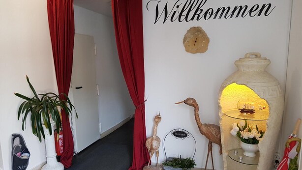 Un pasillo con cortinas rojas, un letrero de bienvenida, un reloj y adornos, incluyendo una escultura de pájaro y un jarrón con flores.