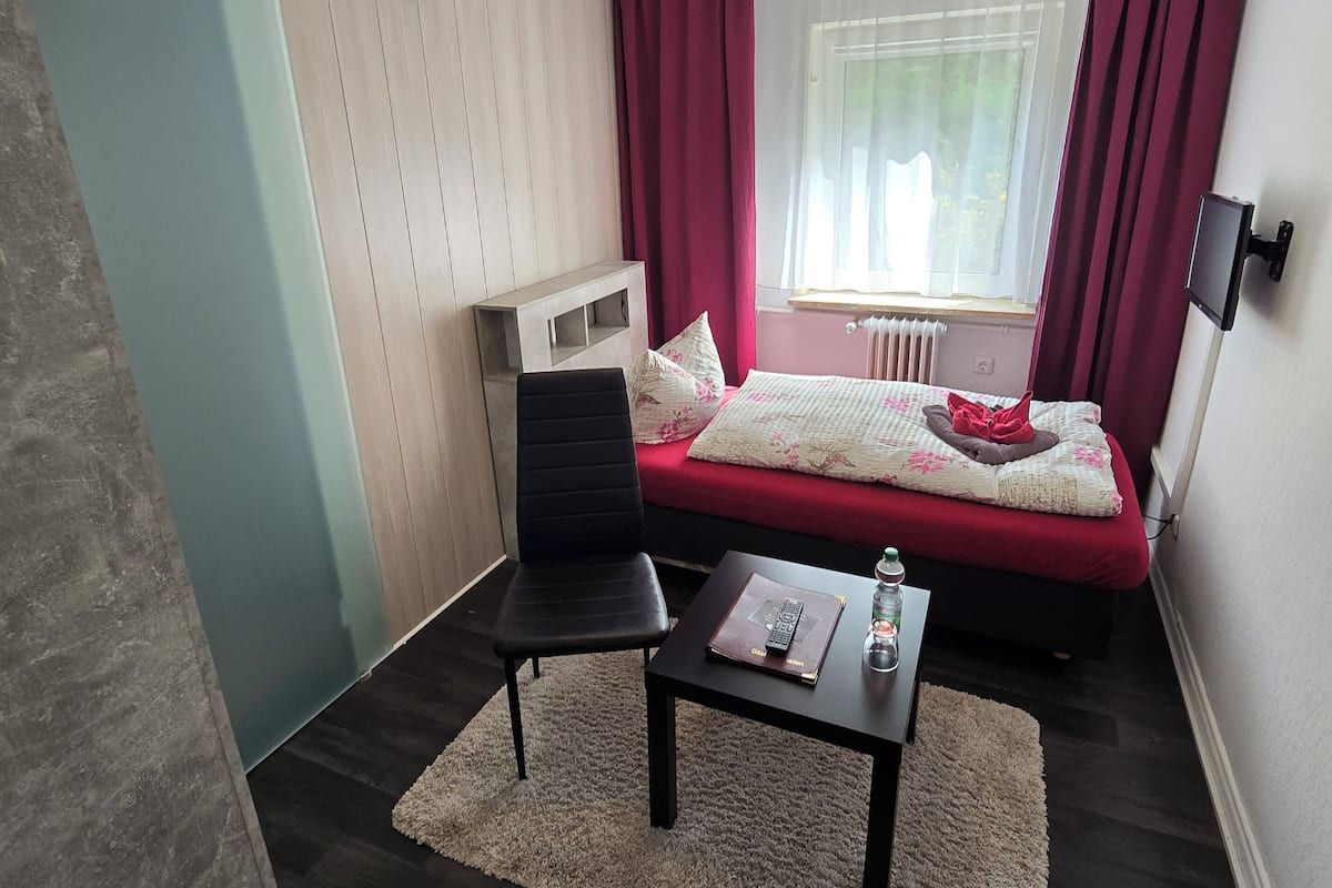 A compact room with a bed, a chair, a small table, and a window with curtains.