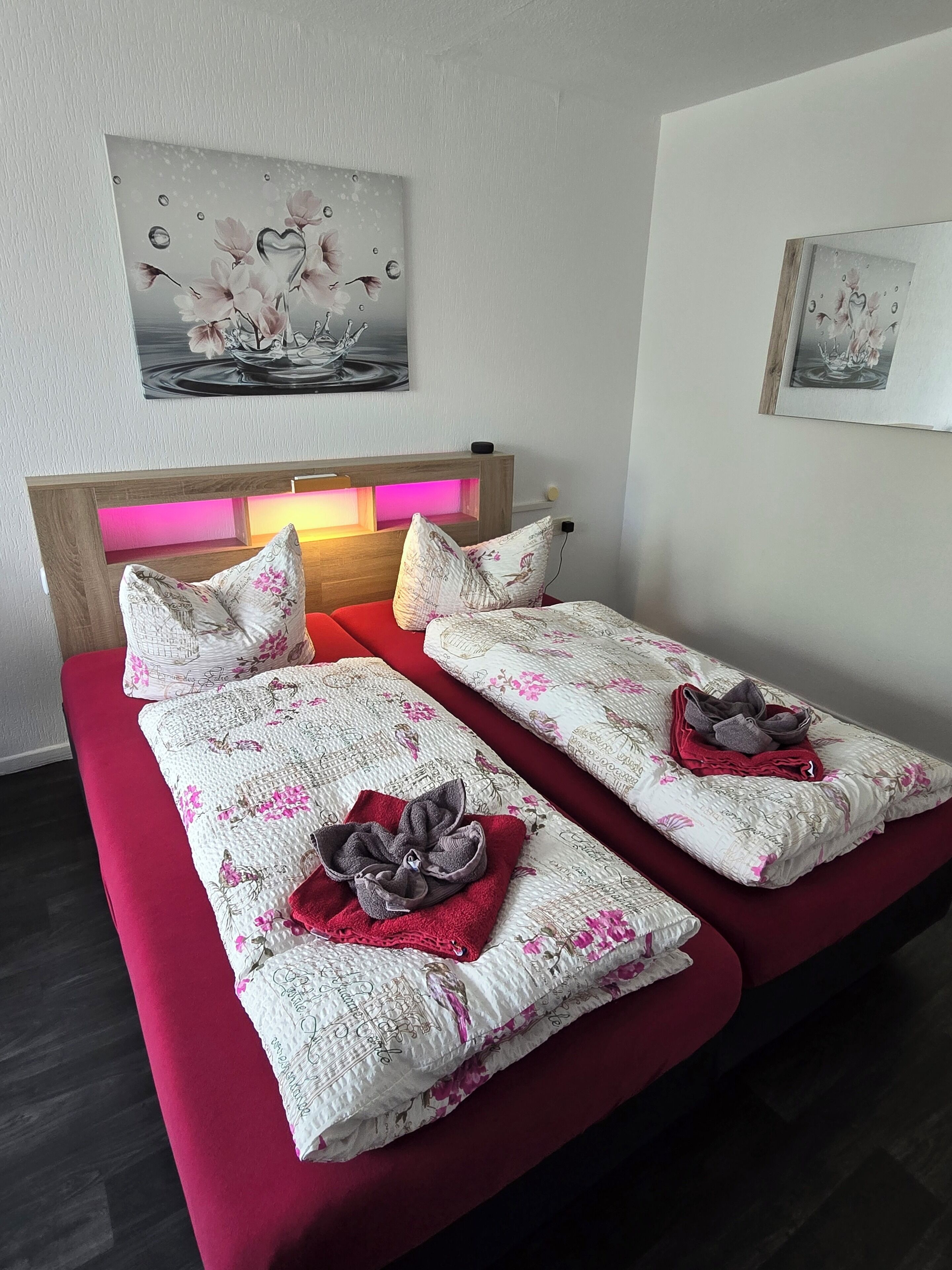 Uma cama de casal com roupa de cama floral e um travesseiro em formato de coração, uma mesinha de cabeceira com abajur e duas obras de arte penduradas na parede.