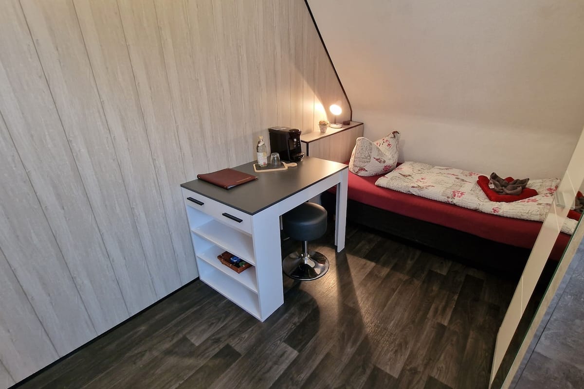A compact room with a desk, a small stool, a bed with a red blanket, and a wall-mounted lamp.