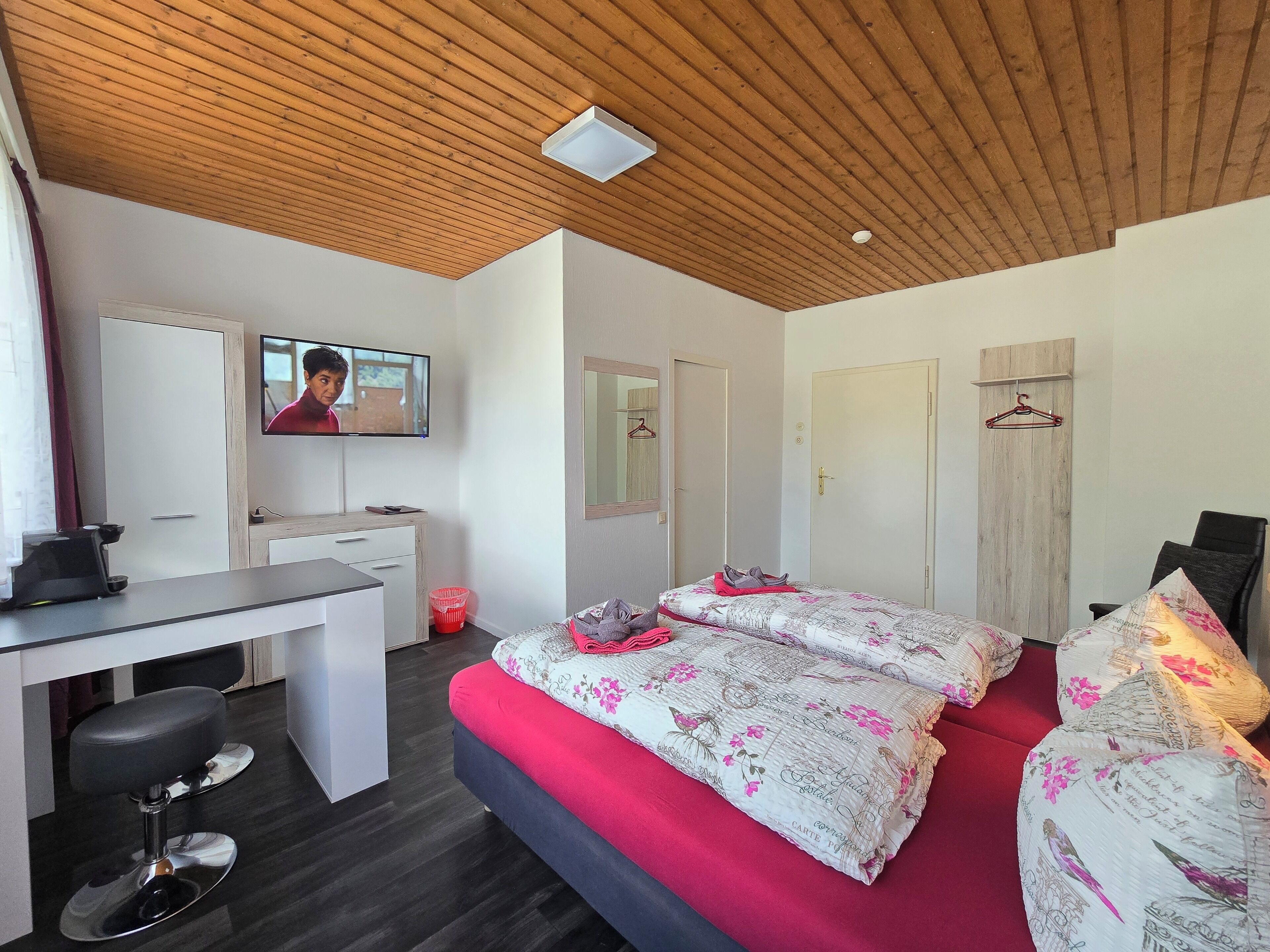 Ein modernes Schlafzimmer mit Holzboden, einem Bett mit floralem Bettzeug, einem Schreibtisch mit Stuhl und einem an der Wand befestigten Fernseher.