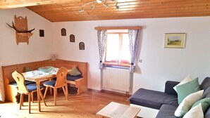 A cozy living room with a wooden ceiling, a dining table with chairs, a sofa with cushions, and a window with curtains. - Ferienwohnung° II 45 qm, extra Schlafzimmer und Ost-Balkon° (Aschau im Chiemgau)