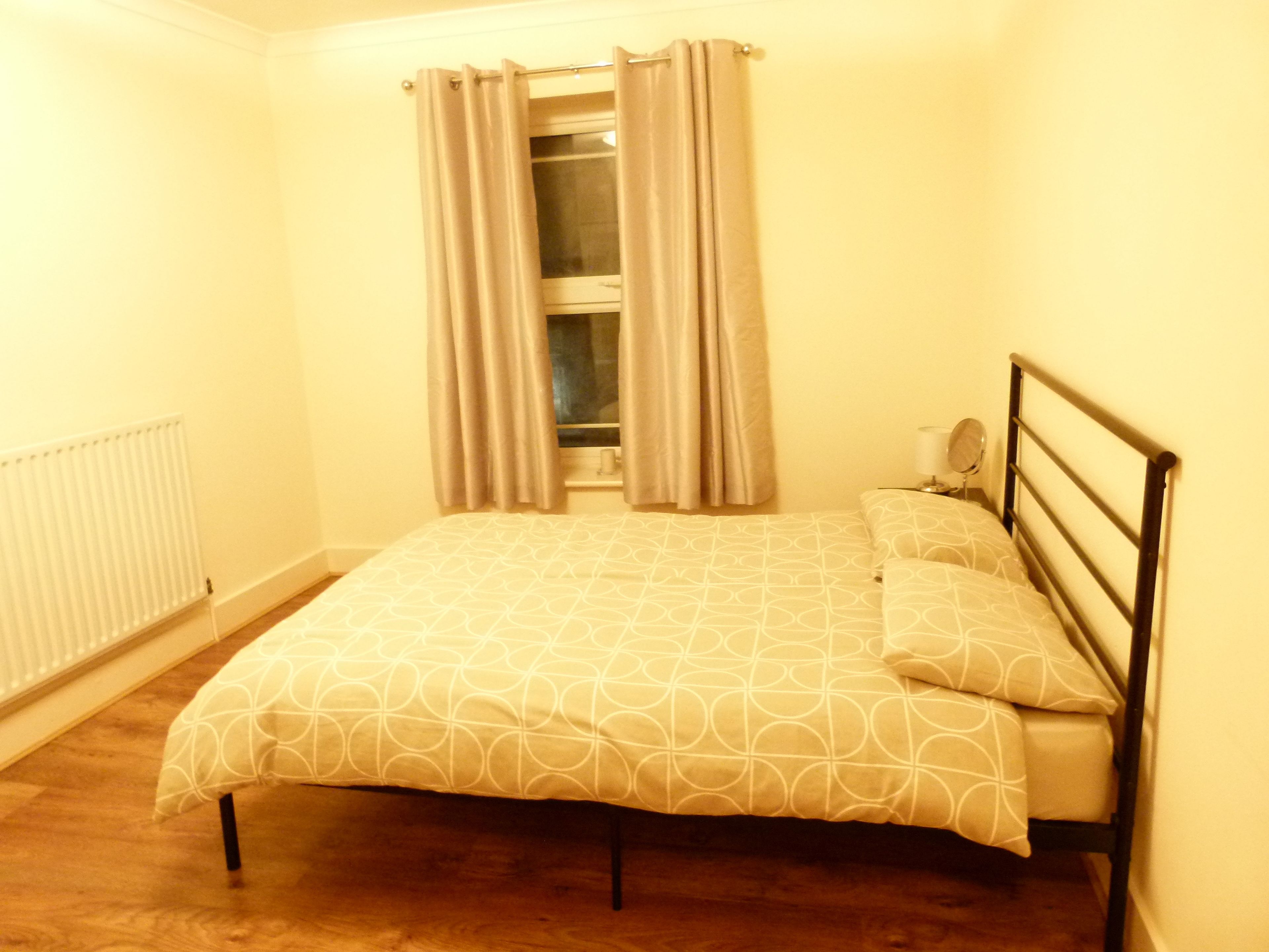 A small, single-bed room with a wooden floor, a radiator, a window with curtains, and a bedside table with a lamp.
