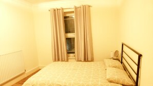 Ein kleines Zimmer mit einem Einzelbett, Holzboden, Heizkörper, einem Fenster mit Vorhängen und einem Nachttisch mit Lampe.