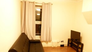 Ein Wohnzimmer mit einer schwarzen Couch, einem Fernseher auf einem Schrank, einem Fenster mit Vorhängen und einem Holzboden.