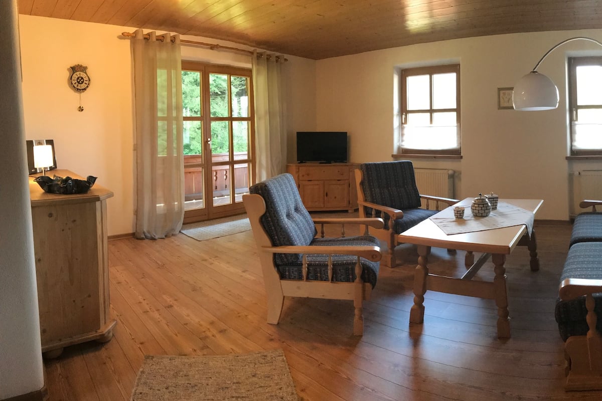 A cozy room with wooden flooring, a sofa, two chairs, a table, a TV, and a lamp.