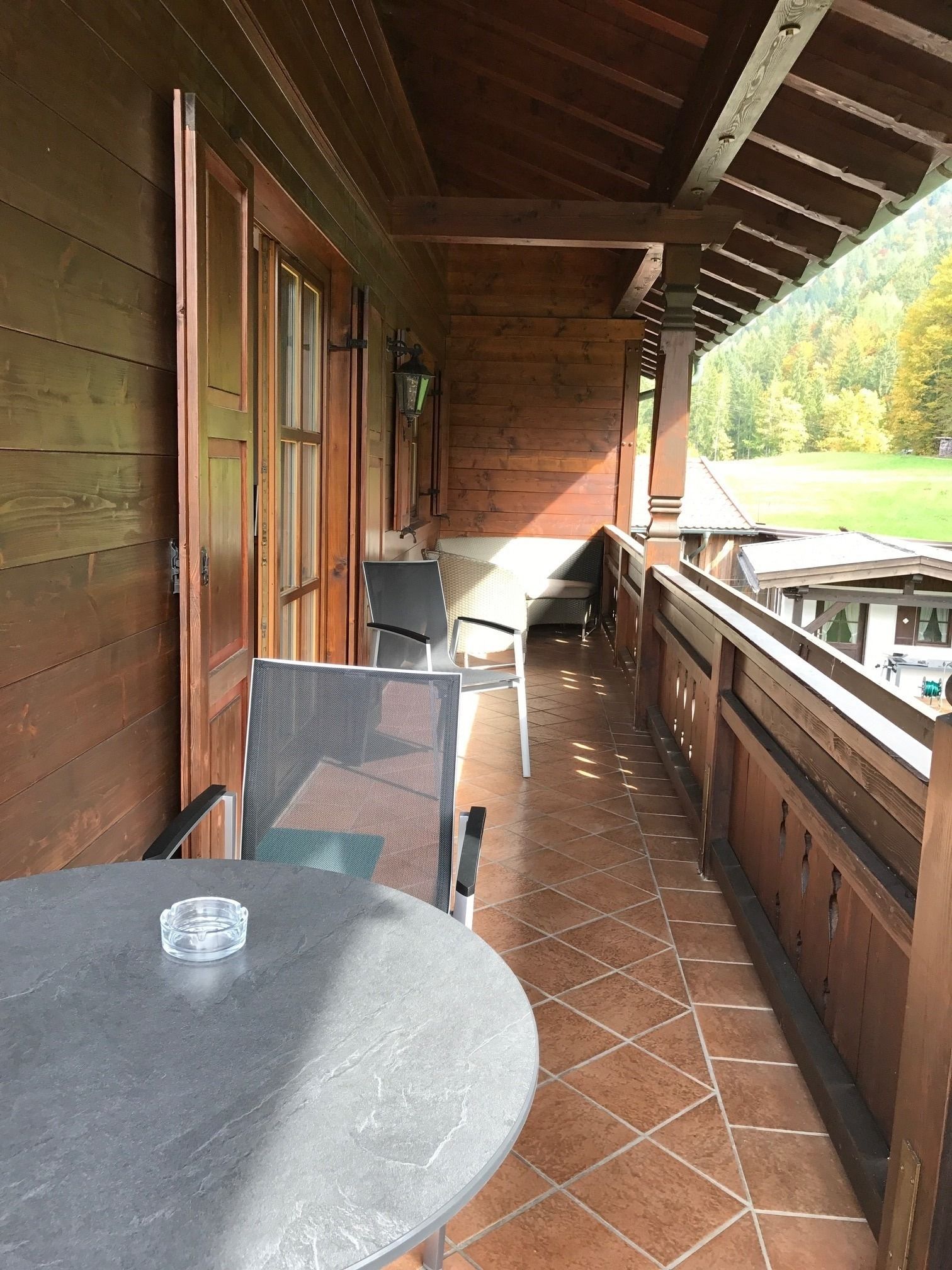 Eine Holzterrasse mit rundem Tisch und Stühlen, Blick auf ein grünes Feld und ein Gebäude mit Holzverkleidung.