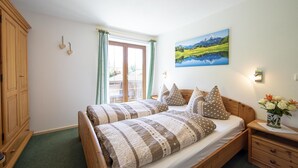 Un dormitorio con una cama de madera, alfombra verde, ventana con cortinas, un cuadro de un paisaje y una mesita de noche con un florero.