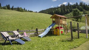 Un parque infantil con un tobogán y una casita, rodeado por una valla de madera y césped.