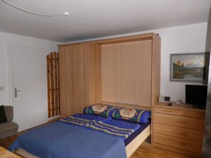 A bedroom with a bed, wooden wardrobe, a desk, and a painting on the wall.
