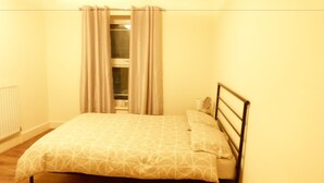 Una habitación pequeña con cama individual, suelo de madera, cabecero de metal y ventana con cortinas.